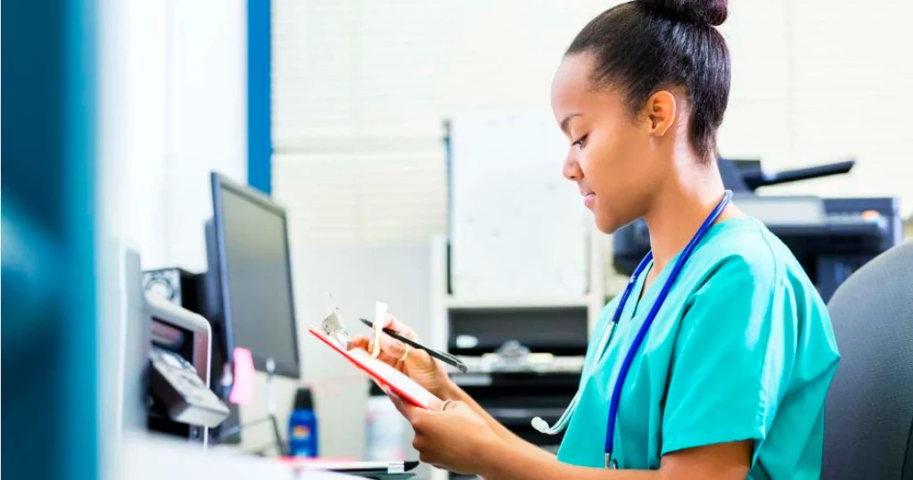 Healthcare professional filling out paperwork and charting at a computer with head bent forward, illustrating tech neck and repetitive strain in health care jobs.