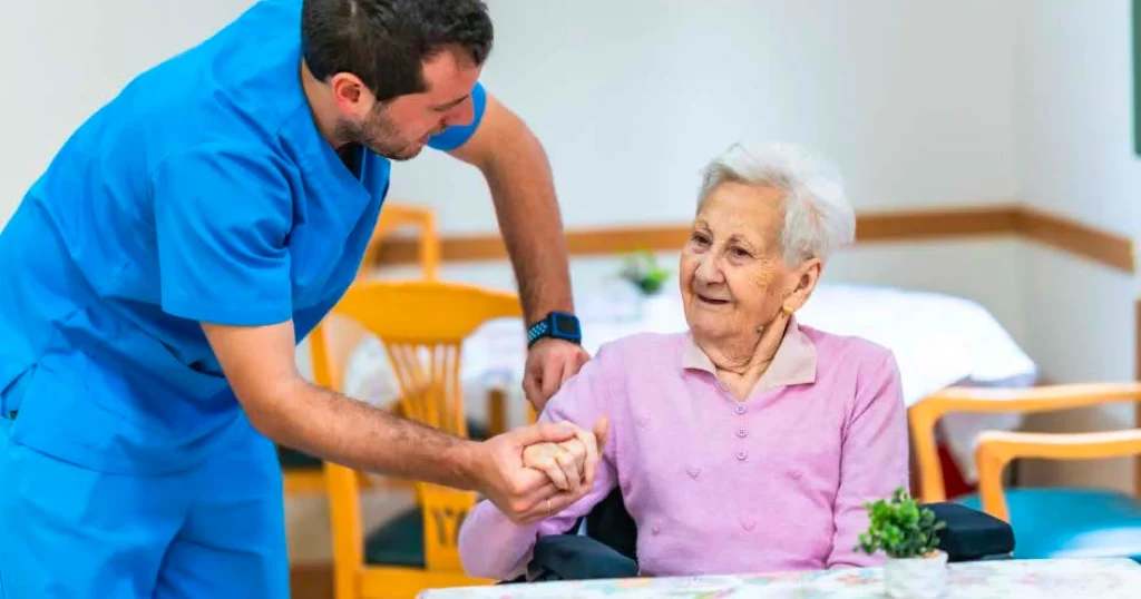 Healthcare professional bending forward to assist an elderly patient in a wheelchair, showing posture strain and the physical demands of daily patient care.