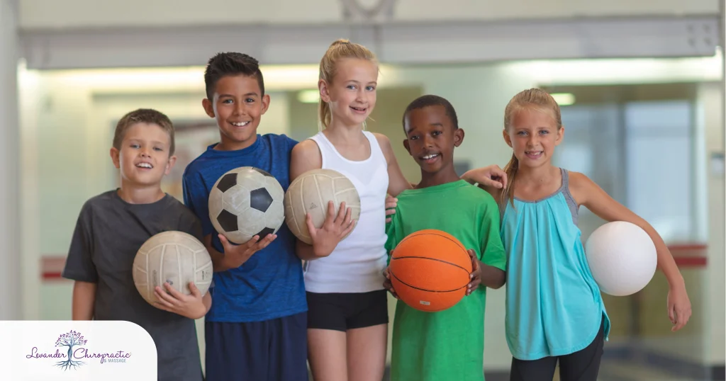 Group of five children standing together in a gymnasium, smiling and holding various sports balls including soccer, volleyball, basketball, and playground balls. The image highlights youth fitness, team sports, and active lifestyles.