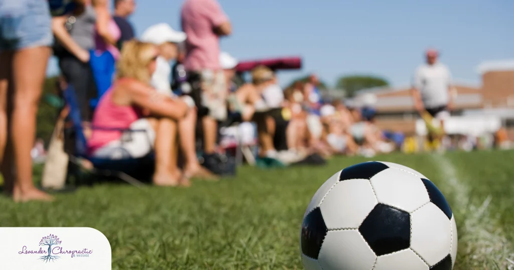 Soccer ball resting on the sideline of a grassy sports field with parents and spectators watching a youth soccer game in the background. The image represents youth athletics, travel sports, and the active lifestyle of sports families.