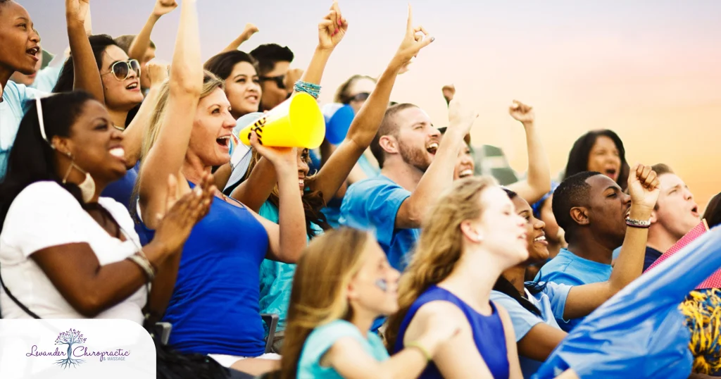 A diverse crowd of enthusiastic fans cheering in stadium bleachers, many wearing blue, with arms raised and mouths open in excitement. One woman holds a yellow megaphone. The Levander Chiropractic & Massage logo appears in the lower left corner.