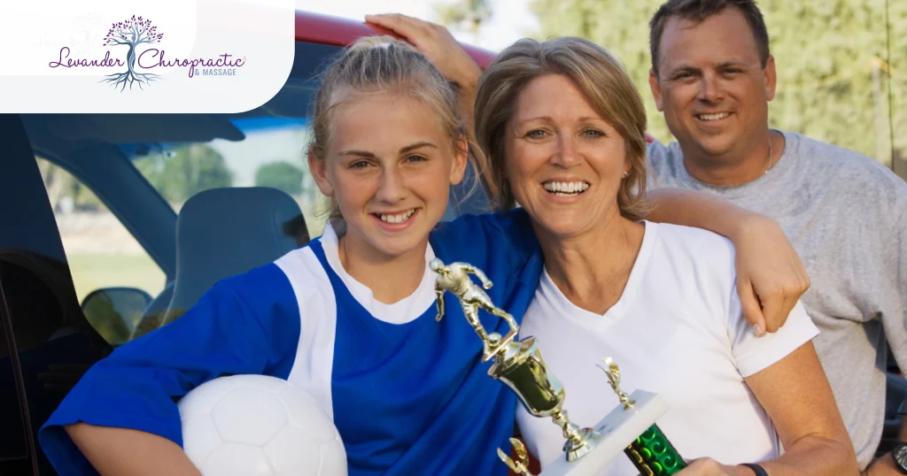 Smiling family standing beside a vehicle after a youth soccer game. A young soccer player in a blue uniform holds a soccer ball and trophy while posing with her parents.