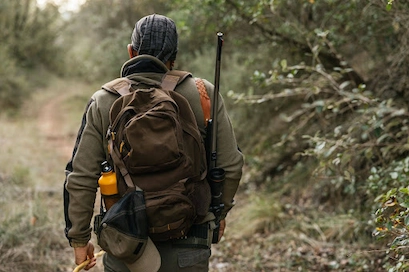 a hunter walks through the woods while carrying a hunting backpack and rifle