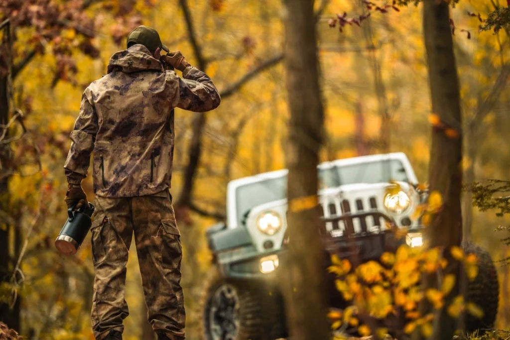 Hunter in camouflage using binoculars in a fall forest, with two off-road vehicles in the background.