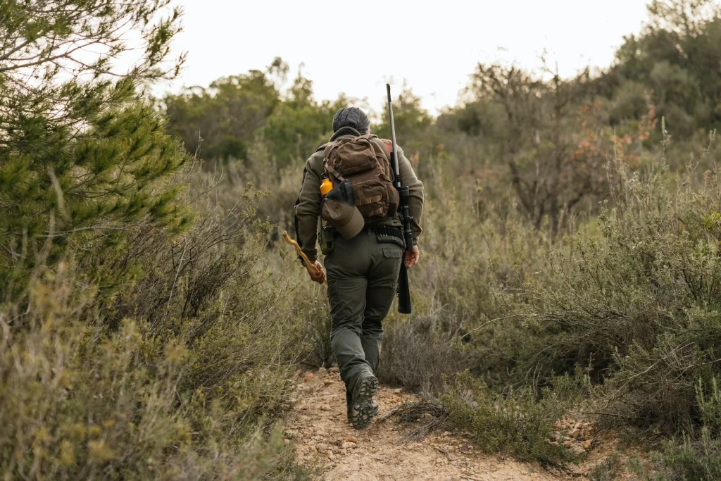 hunter hiking through brush with a rifle and backpack in a wooded area.