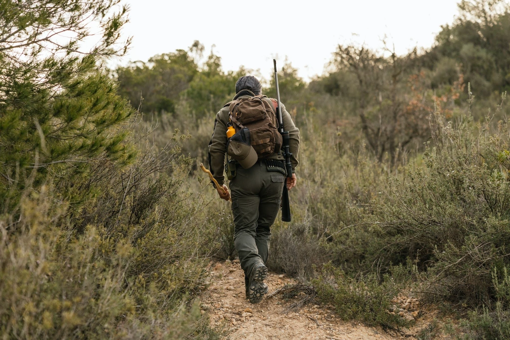 hunter hiking through brush with a rifle and backpack in a wooded area.